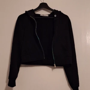 Svart croppad hoodie med dragkedja - Svart croppad hoodie från FILA med huva och dragkedja framtill. Tröjan har långa ärmar och ribbade muddar vid ärmslut och nederkant. Perfekt för dig som gillar en chill och sportig stil. Passar till jeans eller mjukisbyxor.