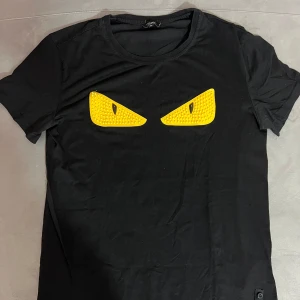 Svart Fendi t-shirt med gula ögon - Cool svart t-shirt från Fendi med två stora gula ögon framtill som ger en edgy vibe. Materialet känns mjukt och skönt mot huden.