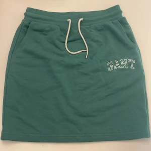 Grön kortkjol med snörning från GANT - Snygg grön kortkjol från GANT med vit snörning i midjan och fickor på sidorna. Kjolen har en sportig vibe och är tillverkad i mjukt bomullsmaterial. GANT-loggan är broderad framtill för en clean look.