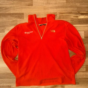 Orange fleece-tröja från The North Face - Säljer en orange fleece-tröja från The North Face i storlek L. Tröjan har half zip, lång ärm och broderad logga på bröstet. Materialet är mjuk Polartec polyester, perfekt för kyliga dagar. Snygg och sportig med Canal Digital-tryck på ena sidan. Mått: längd: 65cm. Bredd: 50cm. Hör av dig vid frågor eller funderingar.