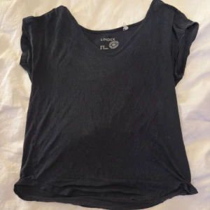 Svart v-ringad t-shirt från Lindex - Svart t-shirt från Lindex med v-ringad hals och korta ärmar. Enkel och stilren design, perfekt för att matcha med jeans eller shorts. Mjuk bomullskvalitet som känns skön mot huden. Passformen är normal och t-shirten har en klassisk look.