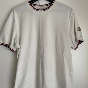 Vit Moncler t-shirt med logga  - Vit t-shirt från Moncler i bomull med klassisk rund hals och korta ärmar. Röd, vit och marinblå rand på krage och ärmslut ger en clean vibe. Moncler-logga på vänster ärm. Perfekt till jeans eller shorts för en chill look.