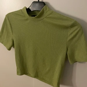 Grön ribbad croppad topp - Snygg grön croppad topp med korta ärmar och ribbad struktur. Toppen har en lite högre hals och är tillverkad i ett stretchigt material som sitter skönt. Perfekt att matcha med jeans eller kjol för en trendig look.