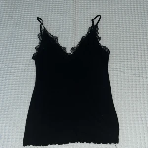 Svart spetslinnetopp med smala axelband - Snygg svart linne-topp med smala axelband och v-ringning. Toppen har fina spetsdetaljer längs urringningen och runt axelbanden, vilket ger en extra touch. Perfekt att styla med jeans eller kjol för en trendig look.