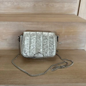 Glittrig silver axelväska med kedja Zara - Säljer en glittrig axelväska i silver från Zara med cool struktur och kedjerem. Väskan har ett lock med magnetknäppning och ett rymligt fack inuti. 