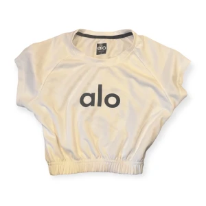 Vit croppad topp från Alo - Snygg vit croppad topp från Alo med svart logga framtill. Toppen har korta ärmar och elastisk midja som ger en cool och sportig vibe. Perfekt för dig som gillar att sticka ut på gymmet eller bara vill ha en chill look.