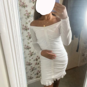 Vit offshoulder kortklänning med volang - Säljer en vit kortklänning med offshoulder och långa ärmar. Klänningen har rynkade detaljer framtill och en söt volangkant nertill. Sitter tight och har en feminin siluett, perfekt för dig som vill sticka ut lite extra.