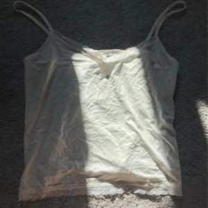 Vit linnetopp med spets från Nelly - Supersöt vit linnetopp från Nelly i storlek XL. Tunn och mjuk bomull med smala axelband och v-ringad hals. Spetsdetaljer längs halsringning och nederkant 