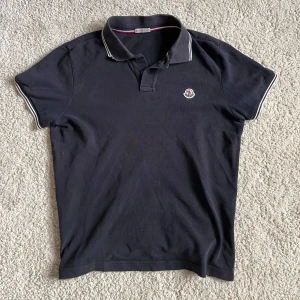 Mörkblå pikétröja från Moncler - Mörkblå pikétröja från Moncler med klassisk krage och korta ärmar. Tröjan har vit piping på krage och ärmslut samt Moncler-logga på bröstet. Tillverkad i mjuk bomull som är skön mot huden. Perfekt för dig som gillar stilrena och sportiga plagg.