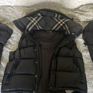 Svart dunväst från Burberry - Säljer en svart äkta  dunväst från Burberry med avtagbara ärmar. Västen har dragkedja och knappar framtill, två fickor med dragkedja och klassiskt rutigt foder i huvan. Perfekt för lager-på-lager och riktigt snygg streetstil. 
