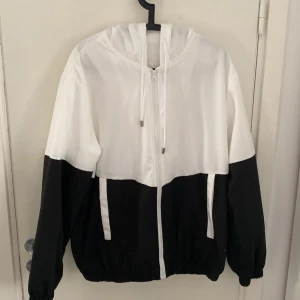Svartvit vindjacka med huva - Snygg svartvit vindjacka med dragkedja och huva. Jackan har elastiska muddar, snörning i huvan och två fickor framtill. Perfekt för blåsiga dagar och enkel att matcha med det mesta. Färgblockad design ger en clean och modern look.