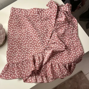 Rosa omlottkjol med volang och blommor - Supersöt kort omlottkjol i ljusrosa med vita små blommor. Kjolen har volangkant och knyts i midjan med en rosett samt knappar, använd enstaka gång