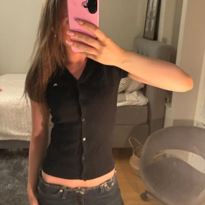 Svart kortärmad topp J.Lindeberg - Svart, figurnära topp från J.Lindeberg i storlek XS. Modellen har korta ärmar, v-ringning och knappar hela vägen framtill. Jätte fin!💞