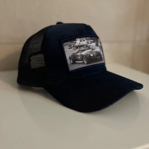 Mörk blå keps med bilmotiv - Snygg blå keps med mesh på sidorna och ett coolt svartvitt tryck av en sportbil framtill. Skärmen är böjd och kepsen har klassisk trucker-stil. Perfekt för dig som gillar bilar och vill ha en unik accessoar.
