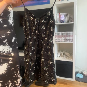 Svart blommig kortklänning med smala axelband - Svart kortklänning med smala axelband och v-ringning, täckt av små blommiga mönster i vitt och rosa. Klänningen har en knyt-detalj framtill och är gjord i ett lätt material som passar perfekt för varma dagar.