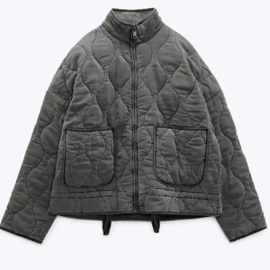 Grå quiltad pufferjacka med dragkedja - Quiltad pufferjacka i mörkgrått med oversized passform och hög krage. Jackan har två stora fickor framtill, dragkedja och coola sömmar som ger en unik look. Perfekt för dig som vill ha en trendig och bekväm jacka till höst och vinter.
