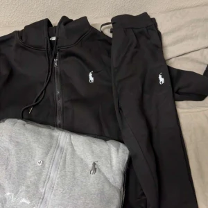 Svart hoodie från Polo Ralph Lauren - Svart hoodie från Polo Ralph Lauren med dragkedja och huva. Klassisk vit logga broderad på bröstet och snörning i huvan. Perfekt för chill dagar eller när du vill ha en clean och enkel look. Materialet är mjukt och skönt, troligtvis bomull eller polyester.