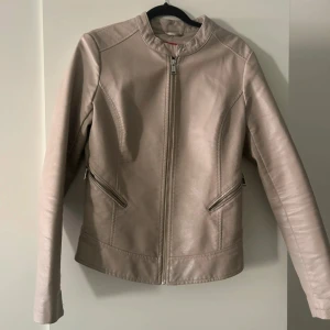 Beige skinnjacka från Guess, stl S - Snygg beige skinnjacka från Guess med stilren design och silvriga dragkedjor. Jackan har två snedställda fickor med dragkedja och en klassisk krage utan slag. Perfekt för dig som vill ha en clean look med edge.