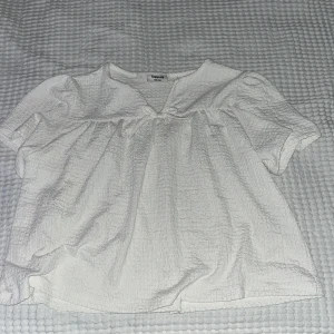 Vit blus med knyt från Kappahl - Supersöt vit blus från Kappahl i skrynklig bomullskänsla. Den har korta ärmar, v-ringning och en liten knytrosett framtill. Blusen är luftig och har en lätt volang under bysten för en avslappnad och trendig look.