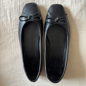 Svarta ballerinaskor från NA-KD - Klassiska svarta ballerinaskor från NA-KD med fyrkantig tå och söt rosettdetalj framtill. Skorna har en stilren design i syntetmaterial och platt sula, perfekta för en clean och chic look.