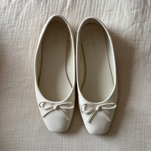 Vita ballerinaskor med rosett från NA-KD - Snygga vita ballerinaskor från NA-KD med fyrkantig tå och dekorativ rosett framtill. Skorna har en stilren och minimalistisk design, perfekt för dig som gillar en clean look. Tillverkade i syntetmaterial och har platt sula för extra komfort.