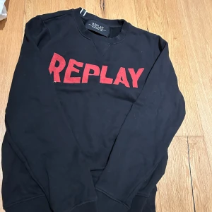 Svart sweatshirt från Replay med röd logga - Svart sweatshirt från Replay i bomull med stor röd REPLAY-logga framtill. Klassisk rund halsringning och långa ärmar. Perfekt för dig som gillar streetwear och vill sticka ut med en tydlig logga.