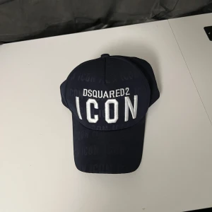 Mörkblå Dsquared2 ICON keps - Snygg mörkblå keps från Dsquared2 med stor vit broderad ICON-text framtill. Justerbar metallspänne med ICON-gravyr baktill. Kepsen har böjd skärm och diskret ton-i-ton ICON-mönster över hela tyget. Perfekt för dig som gillar streetwear och vill sticka ut.