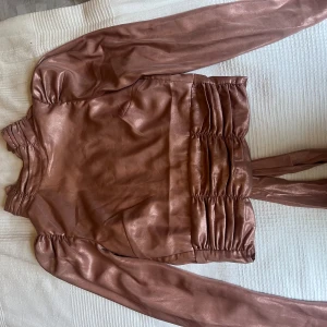 Glansig fest topp med öppen rygg - Säljer en glansig metallrosa omlottopp från Gina Tricot med långa ärmar och breda knytband i midjan. Toppen har lätt puff vid axlarna och smockdetaljer vid ärmslut och midja. Toppen har en öppen rygg med knyte där bak. SKRIV INNAN DU KÖPER!