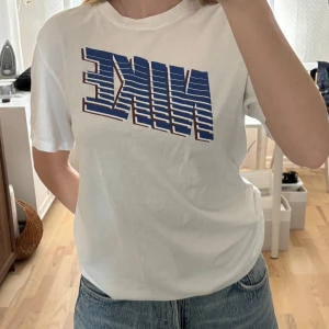 Vit Nike t-shirt med blått tryck - Vit t-shirt från Nike med ett stort, blått och randigt Nike-tryck med vinröda detaljer på bröstet. Klassisk passform. Tillverkad i mjuk bomull som är skön att ha på sig. 