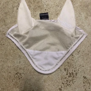 Vit huva från Catago Equestrian - Snygg vit huva från Catago Equestrian med mesh-tyg och broderad logga på ena örat. Huvan har en glittrig kant runtom. Tvättas innan frakt, finns även schabrak att köpa till k hoppmodell också storlek full. 