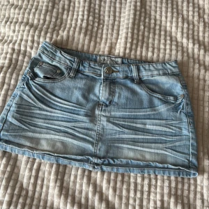 Ljusblå jeanskjol från J&J Jeans - Snygg ljusblå jeanskjol från J&J Jeans i klassisk femficksmodell med knapp och dragkedja framtill. Kjolen har slitningar och tvättade detaljer som ger en cool vibe. Passar perfekt till sneakers eller boots och har bälteshällor för extra styling. Den sitter som ungefär M och S