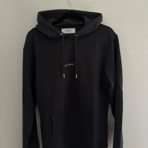 Les Deux hoodie (mörkblå) - Blå hoodie från Les Deux med stilren design och vit broderad logga på bröstet. Tröjan har huva med snörning, stor magficka och långa ärmar. Perfekt för chill dagar och enkel att matcha med jeans eller joggers.