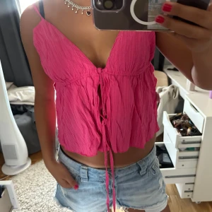 Rosa linnetopp med knyt framtill - Supersöt rosa linnetopp med smala axelband och djup v-ringning. Toppen har en luftig, vågig struktur och knytband framtill som ger en avslappnad och trendig look. Perfekt för varma dagar och enkel att styla med jeansshorts eller kjol.
