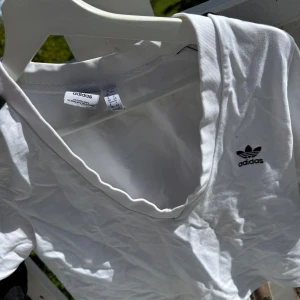 Vit v-ringad topp från Adidas - Vit v-ringad topp från Adidas med klassisk svart logga på bröstet. T-shirten har korta ärmar och är gjord i mjuk bomull, perfekt för en sportig och clean look. Enkel att matcha med jeans eller shorts för en avslappnad stil.