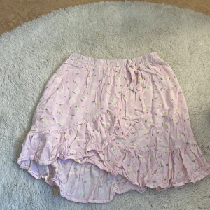 Rosa blommig omlottkjol med volang - Supersöt rosa kortkjol med små vita och gula blommor. Kjolen har omlott-design med knyt i sidan och volang längs nederkanten. Perfekt för sommarens alla dagar, mjukt och lätt material som känns skönt mot huden.