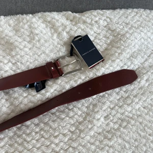 Brunt skärp i skinn från Tommy Hilfiger - Snyggt brunt bälte från Tommy Hilfiger i äkta skinn med klassisk silverfärgad metallspänne. Aldrig använd, nypris ca 600kr. Storlek 85.