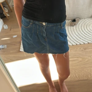 Blå jeanskjol med rå kant - Säljer en blå jeanskjol med rå, fransig kant och klassiska fickor både fram och bak. Kjolen har knapp och dragkedja framtill och är i en kort modell som ger en chill vibe. Perfekt för dig som gillar en enkel och cool stil. Mango 