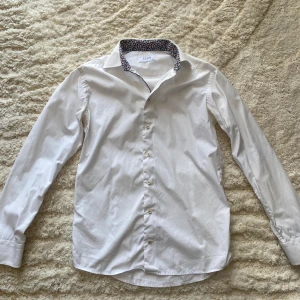 Skjorta från Eton - Snygg vit skjorta från Eton i slim fit med klassisk krage och blommigt mönster på insidan av kragen. Skjortan har långa ärmar och knappar framtill. Perfekt för dig som gillar stilrena plagg med en twist. Materialet är mjuk bomull. Storlek 39.