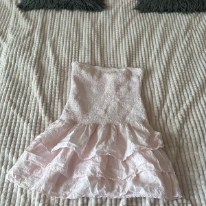 Ljusrosa volangklänning med spets - Supersöt ljusrosa kortklänning med smockad överdel och flera lager volanger i kjolen. Klänningen är axelbandslös och har fina spetsdetaljer med broderade små blommor. Perfekt för sommarens alla tillfällen!