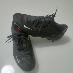 Nike Phantom GT2 Svarta Fotbollsskor - Nike Phantom GT2 fotbollsskor i svart med vita och orange detaljer. Skorna har snörning, mönstrad ovandel och platt sula med orange dobbar för grymt grepp på planen. Snyggt Nike Swoosh på sidan och undersidan. Perfekt för dig som vill ha stil och kontroll på fotbollsplanen.