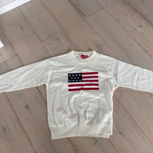 Vit stickad tröja med USA-flagga - Vit stickad tröja från RYVLS i storlek 158/164. Framsidan har en stor USA-flagga i blått, rött och vitt. Tröjan har rund hals, ribbade muddar och lång ärm. Perfekt för dig som gillar clean stil med en cool detalj framtill.
