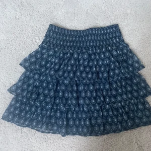 Blå volangkjol från Lindex - Supersöt blå kortkjol från Lindex med vitt mönster och flera volanger. Kjolen har smock i midjan för extra bekväm passform och är perfekt för dig som gillar en luftig och lekfull stil. Tillverkad i mjukt material som känns skönt mot huden.