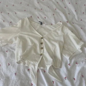 Vit croppad blus från H&M XS - Supersöt vit croppad blus från H&M med v-ringning, knappar framtill och knytband i midjan. Korta ärmar och lätt, luftig bomull gör den perfekt för varma dagar. Enkel men ändå stylish detalj till din outfit.