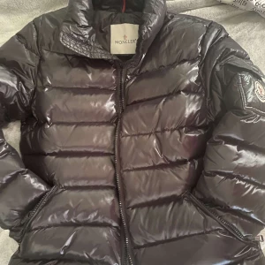 Svart Moncler pufferjacka - Säljer en svart pufferjacka från Moncler med glansig finish och klassisk logga på ärmen. Jackan har dragkedja framtill, hög krage och quiltad design. Perfekt för kalla dagar när du vill ha både stil och värme.