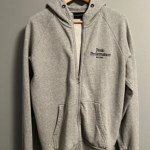 Grå hoodie från Peak Performance - Snygg grå hoodie från Peak Performance med broderad logga på bröstet. Tröjan har huva, dragkedja framtill och två stora fickor. Tillverkad i mjuk bomullsmix, perfekt för chill eller träning. Långärmad och med ribbade muddar för extra komfort.