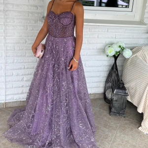 Lila glittrig långklänning med smala axelband - Säljer en magisk lila långklänning med smala axelband och glittriga paljetter över hela klänningen. Överdelen har en snygg korsettlook med markerade sömmar och pärldetaljer. Kjolen är vid och faller snyggt ner mot golvet, perfekt för bal. Klänningen är i storlek 36 men passar även 34