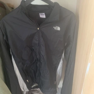 Svart vindjacka från The North Face - Svart vindjacka från The North Face med dragkedja framtill och två fickor med dragkedja. Lätt och smidig modell med klassisk logga på bröstet. Perfekt för blåsiga dagar och enkel att ta med i väskan.