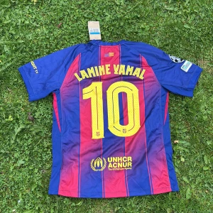 FC Barcelona Lamine Yamal tröja Nike M - Säljer en officiell FC Barcelona matchtröja från Nike med Lamine Yamal och nummer 10 på ryggen. Tröjan är blå och röd med tunna vertikala ränder, gula detaljer och klubbmärke på bröstet. Materialet är lätt polyester med Dri-FIT-teknologi. Spotify-logga på framsidan.