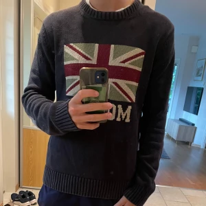 Mörkblå stickad tröja Morris - Snygg mörkblå stickad tröja från Morris med Union Jack-flagga och texten 'MORRIS' på bröstet. Tröjan har rund halsringning, ribbade muddar och är långärmad. Perfekt för dig som gillar klassisk preppy stil.