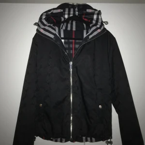 Burberry jacka - Säljer svart burberry jacka. Jackan är dubbelsidig där ena sidan är svart med mönster på hela jackan och knappar för att stänga fickorna, medans den andra sidan är svart med röda och vita ränder med text runt jackan och dragkedja för att stänga fickorna. Jackan är M men passar också bra på S om man vill ha den lite större. Pris kan självklart diskuteras.